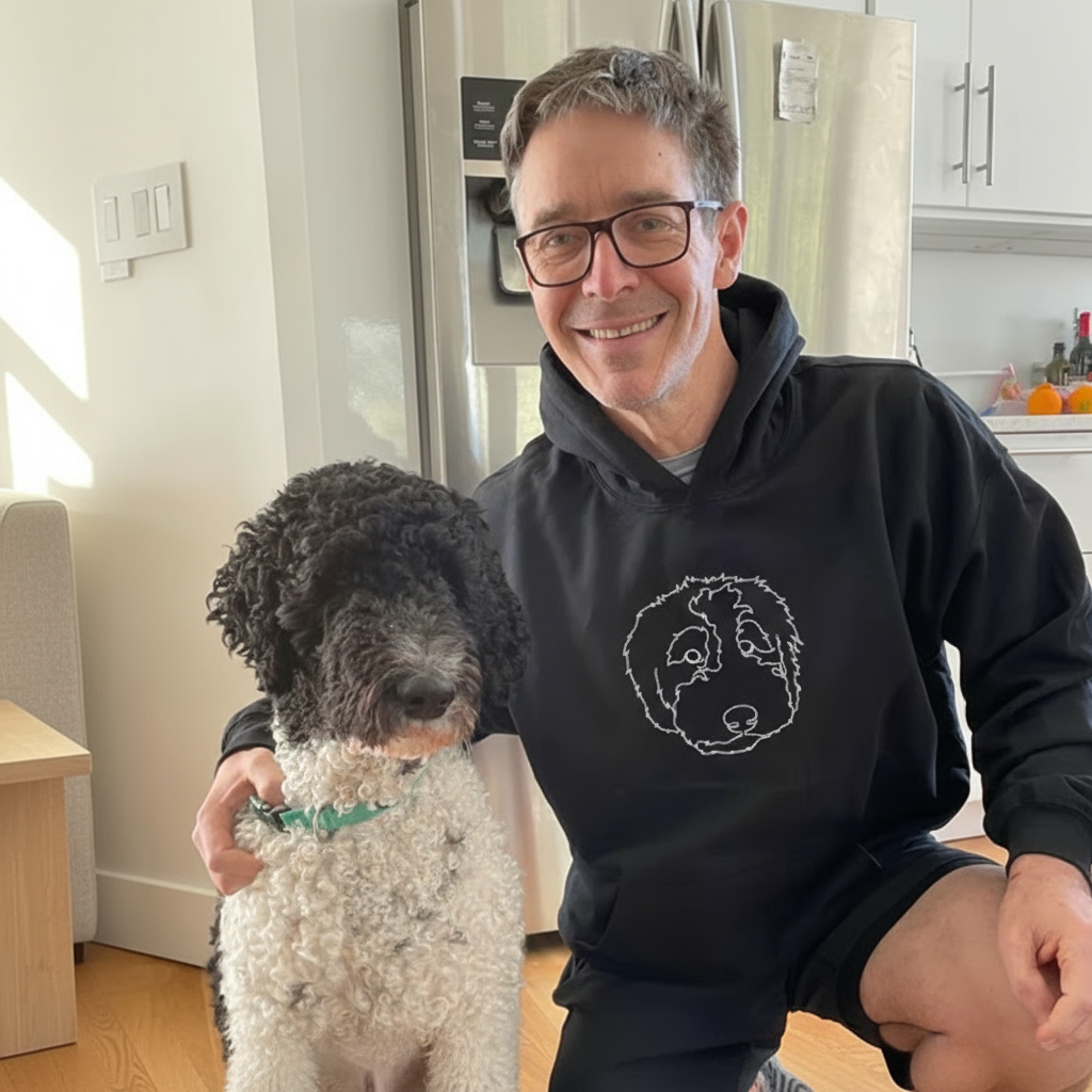 Man wearing a black hoodie with a dog design, sitting with a dog in a kitchen.