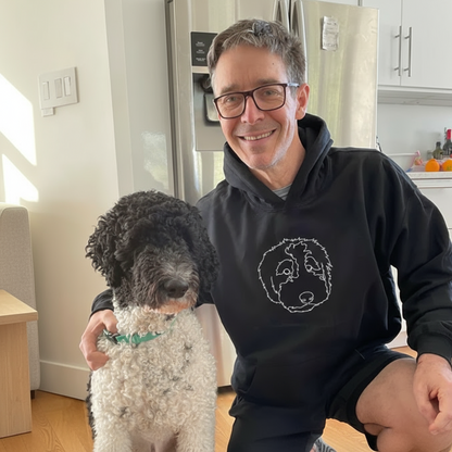 Man wearing a black hoodie with a dog design, sitting with a dog in a kitchen.